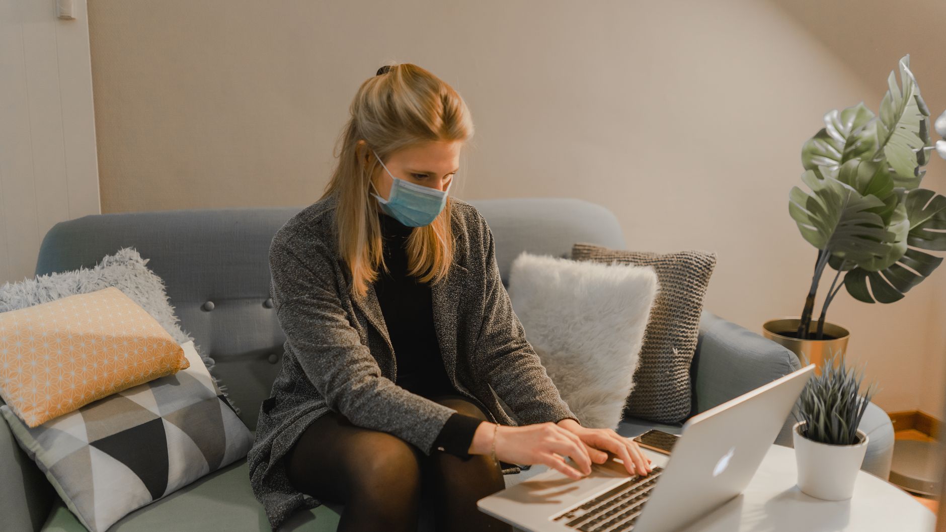 A woman sits on a sofa wearing a face mask while working on a laptop indoors.
