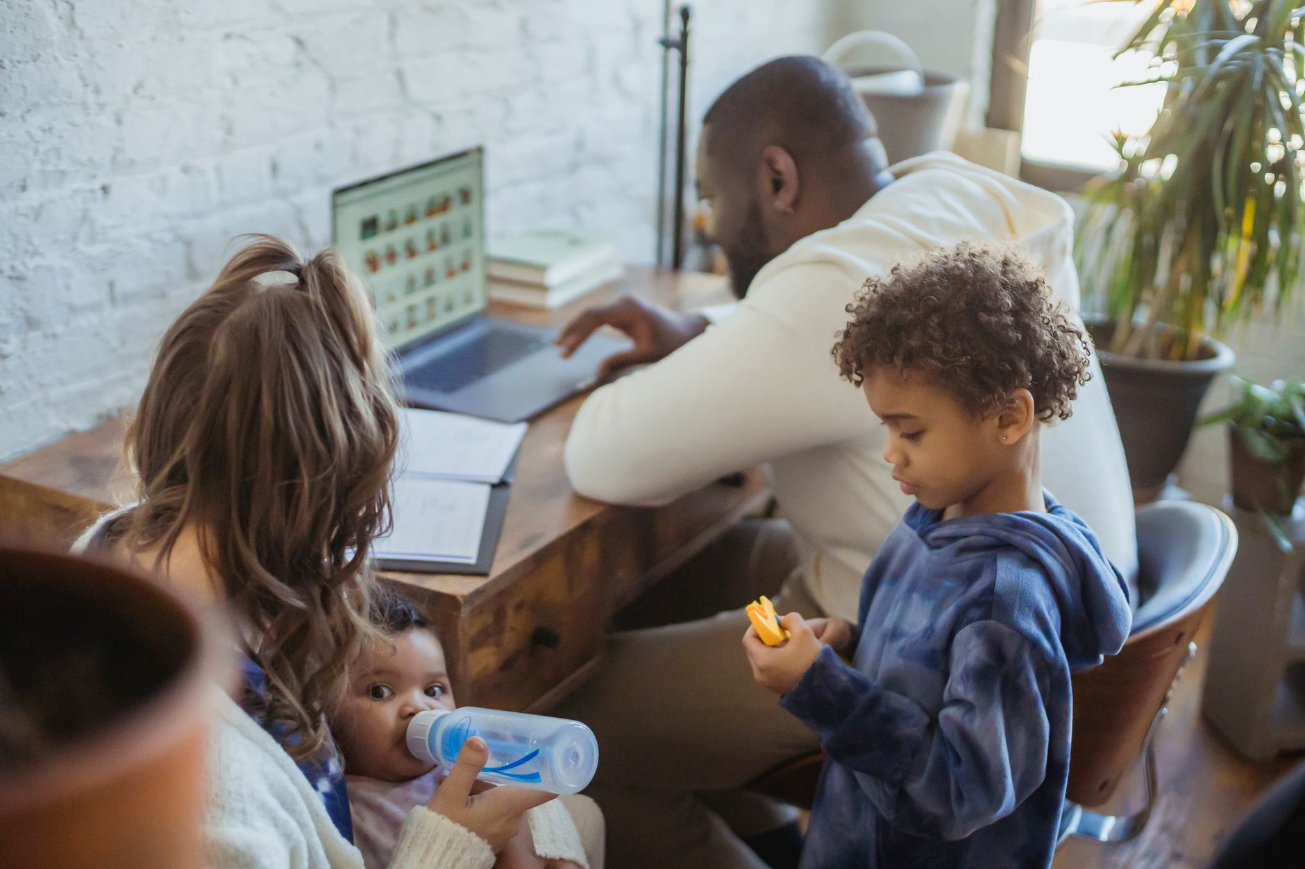 Anonymous mother bottle feeding black baby in living room with African American son and husband browsing modern laptop at home