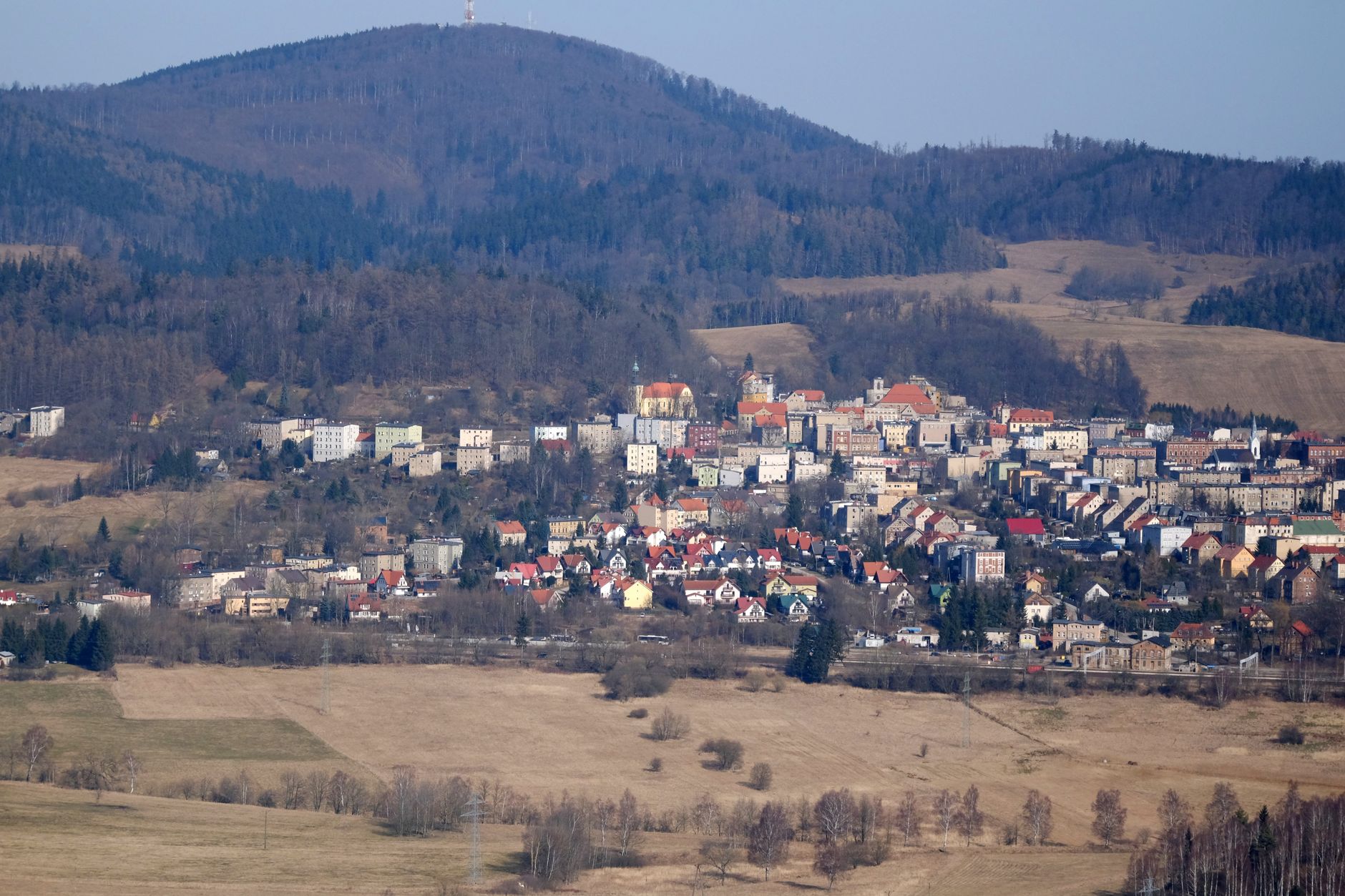 Aerial view of a quaint town nestled among rolling hills and forests, with a mountainous backdrop.
