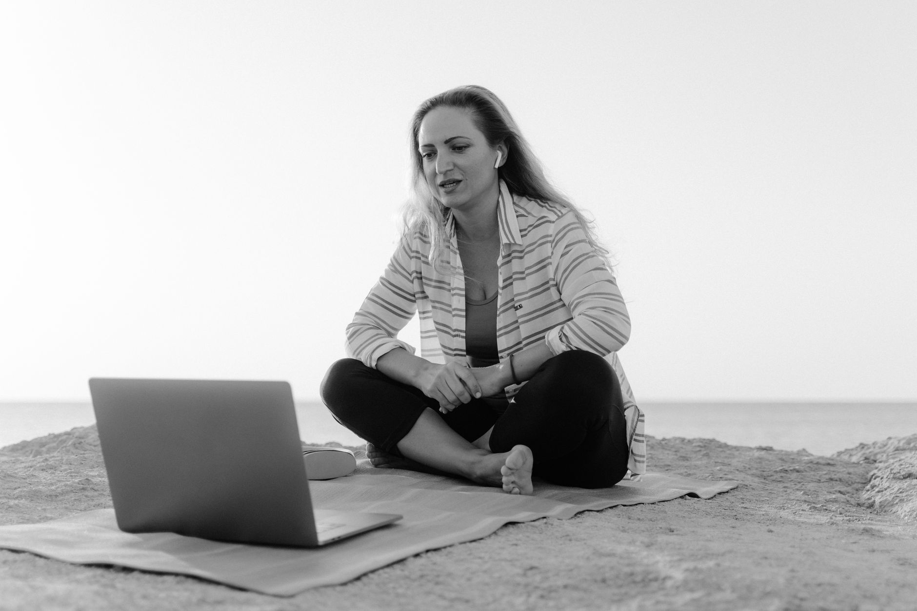 A woman sits on a beach, using a laptop for a video call, offering a peaceful working environment.
