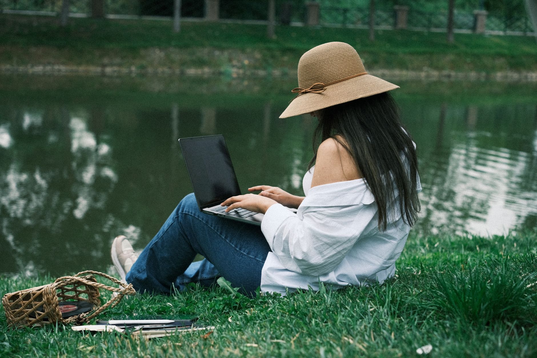 A woman sits by a lake, working on her laptop in a serene park setting.