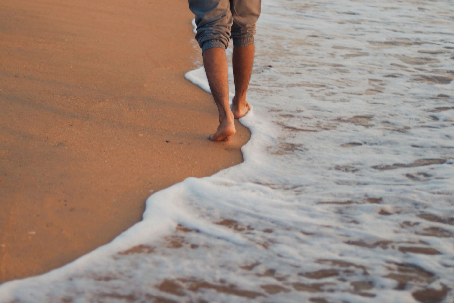 A person walks barefoot along a sandy beach with gentle waves at sunrise, capturing a peaceful moment.
