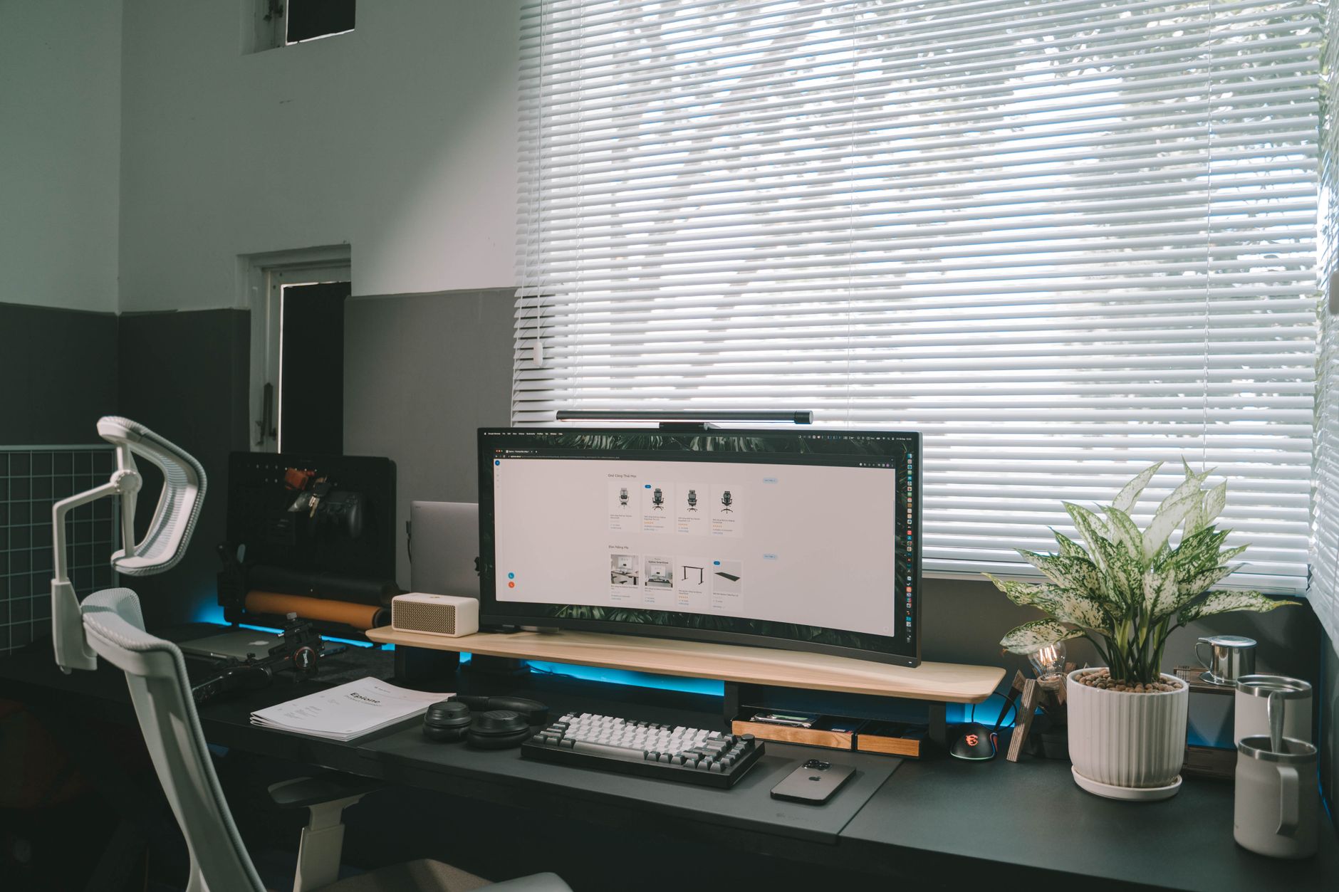 A modern office desk setup featuring a computer monitor, ergonomic chair, and potted plant by a window.