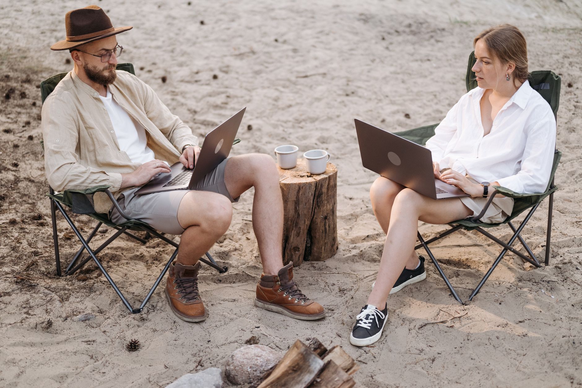 A couple enjoying remote work in nature, using laptops at a sandy campsite, embracing a digital nomad lifestyle.