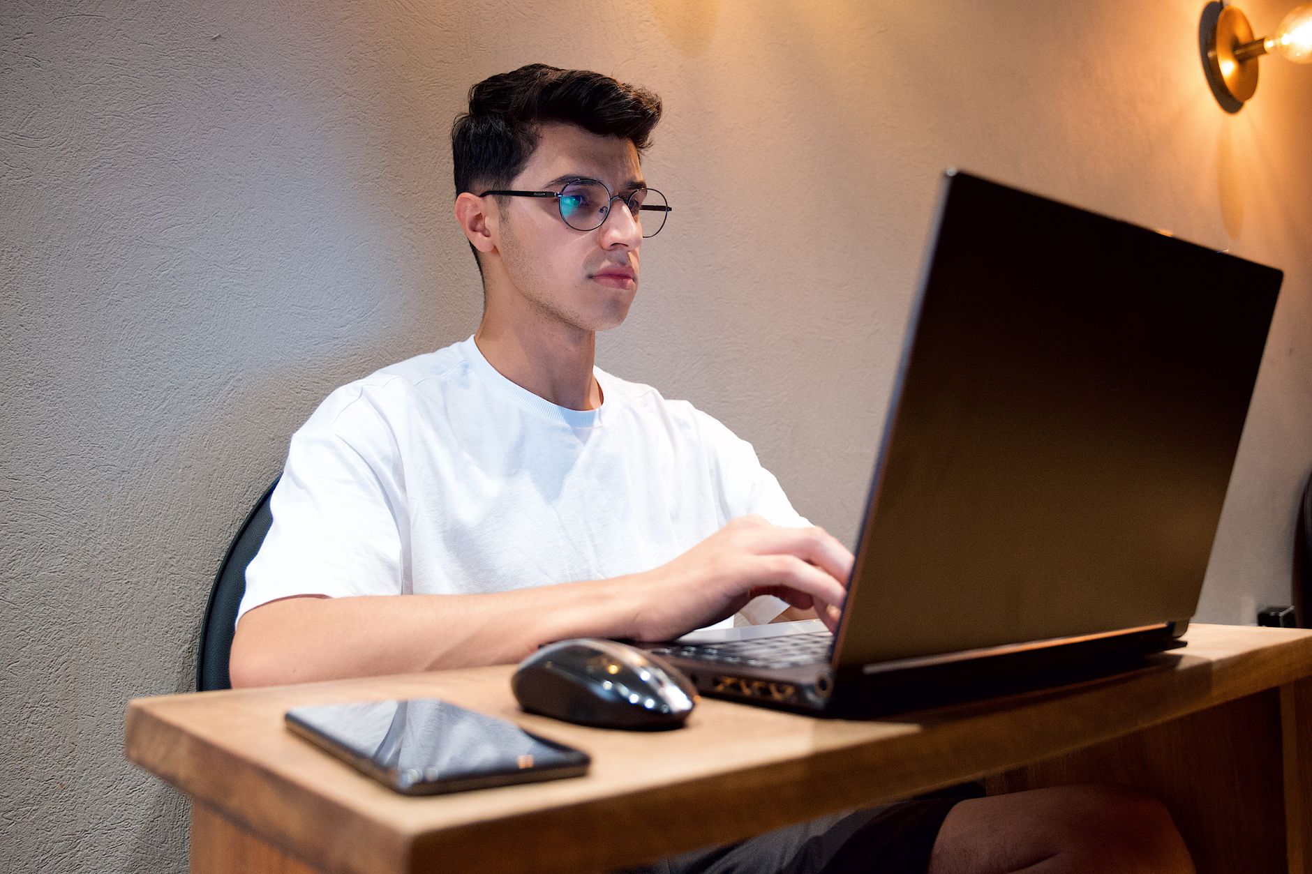 Man in white shirt using laptop at desk indoors with smartphone and mouse nearby.