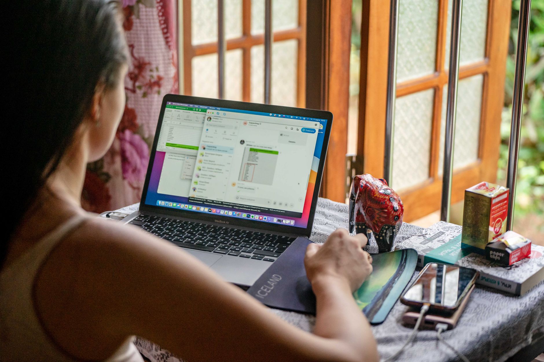 Woman working remotely with a laptop and smartphone in a cozy room near a window.