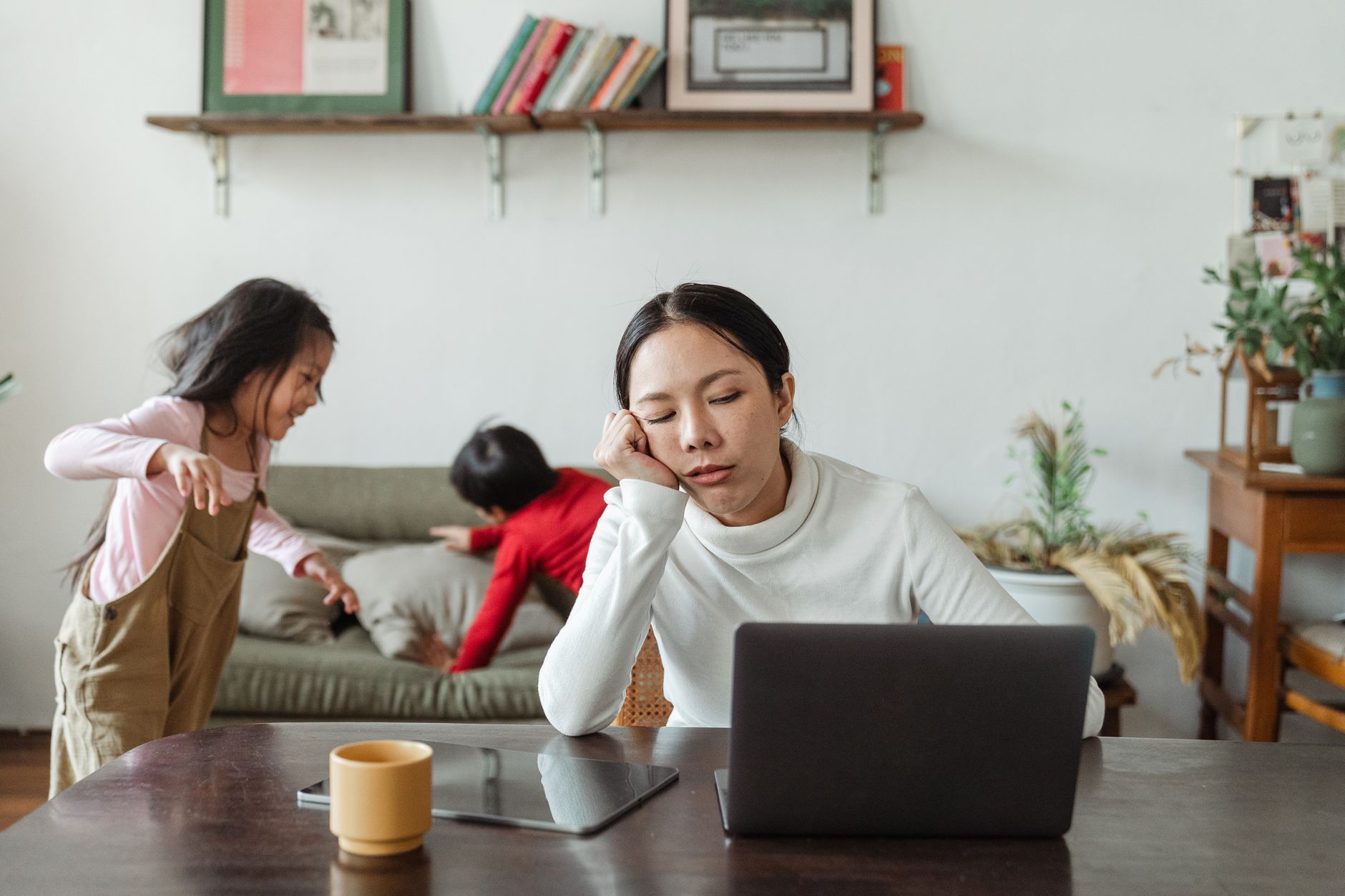 Overworked young Asian female freelancer sitting at table with laptop and tablet against active disturbing children