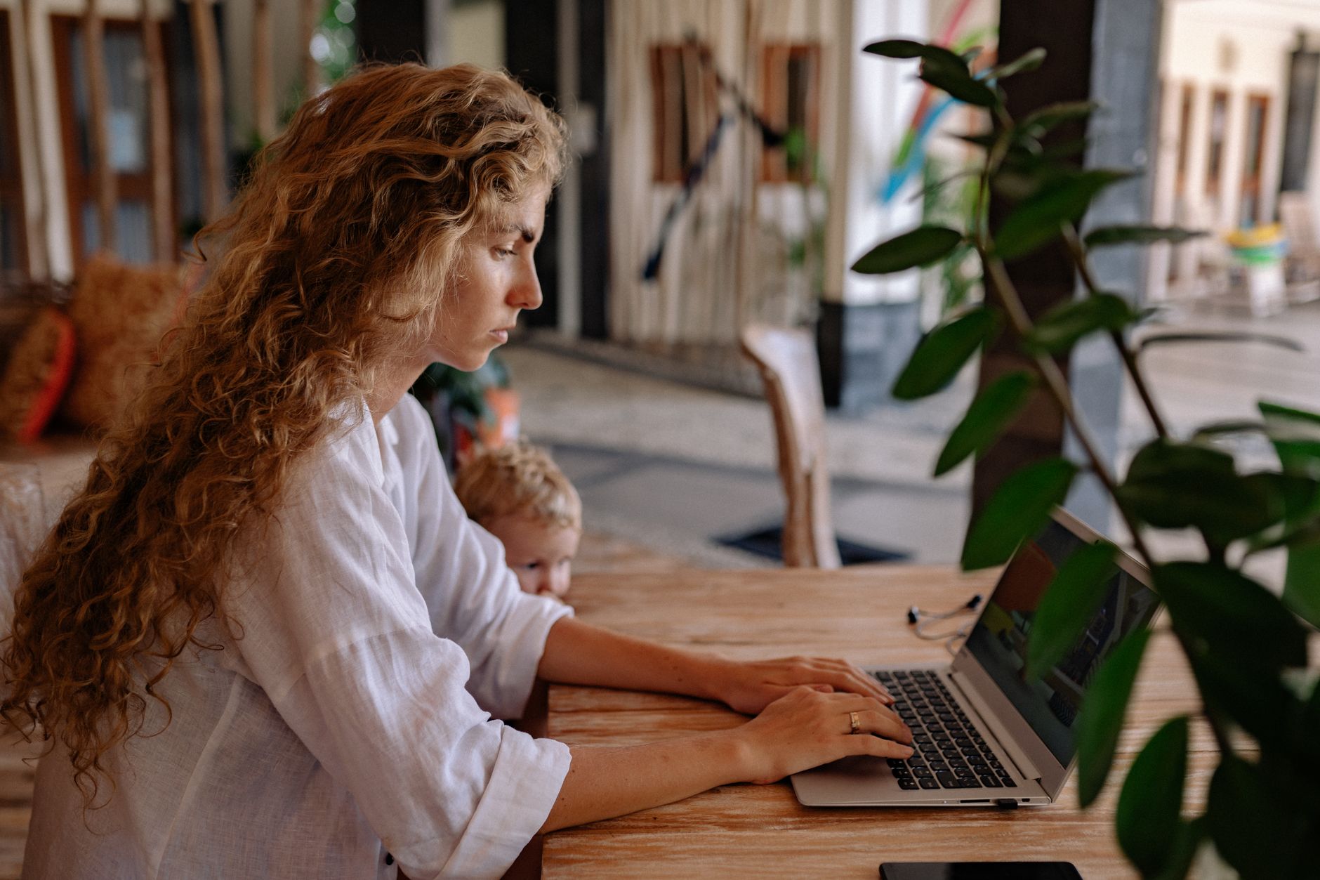 Woman with curly hair working on a laptop at home, with a child nearby, showcasing remote work lifestyle.