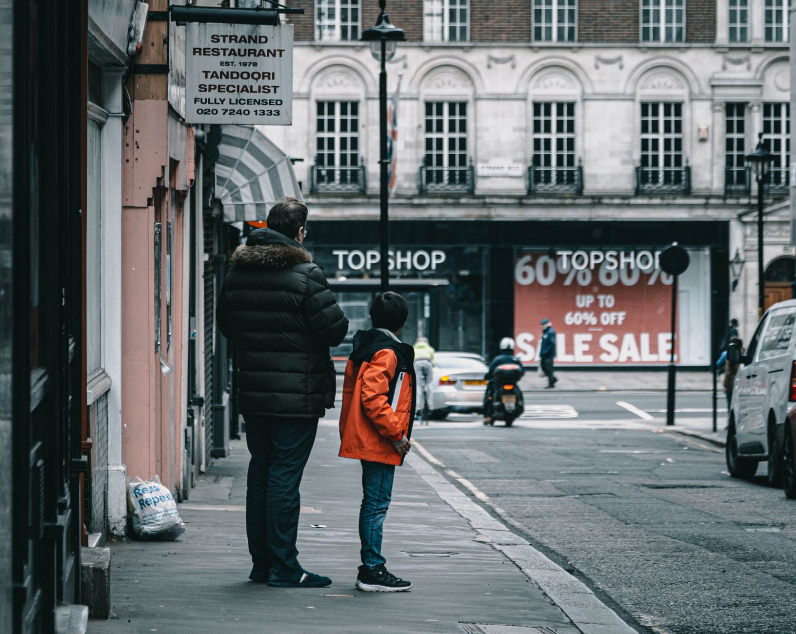 Father and son waiting on a London sidewalk near Strand Restaurant and Topshop signage.